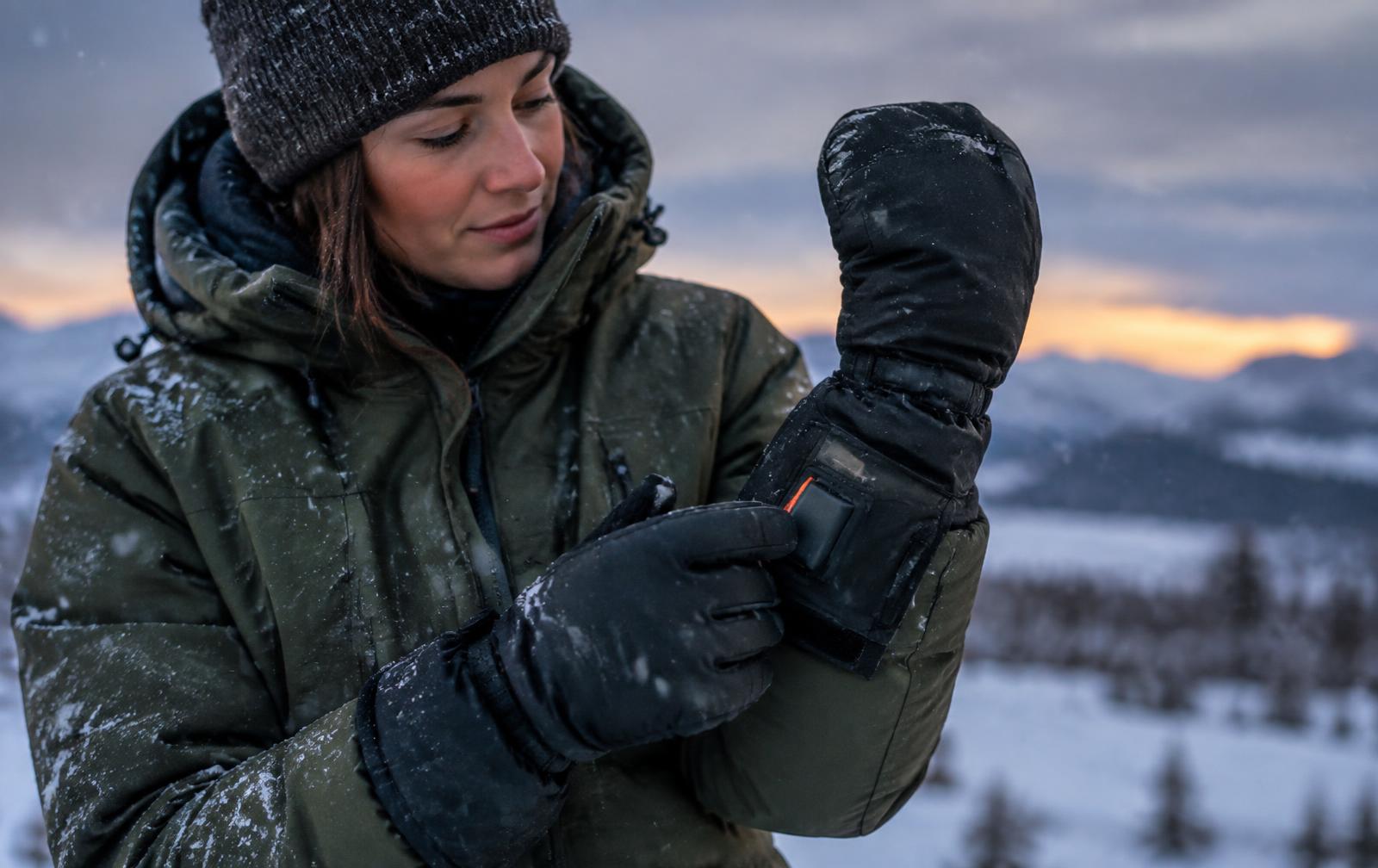 A woman in a winter parka adjusting the battery pocket on a heated mitten at dusk