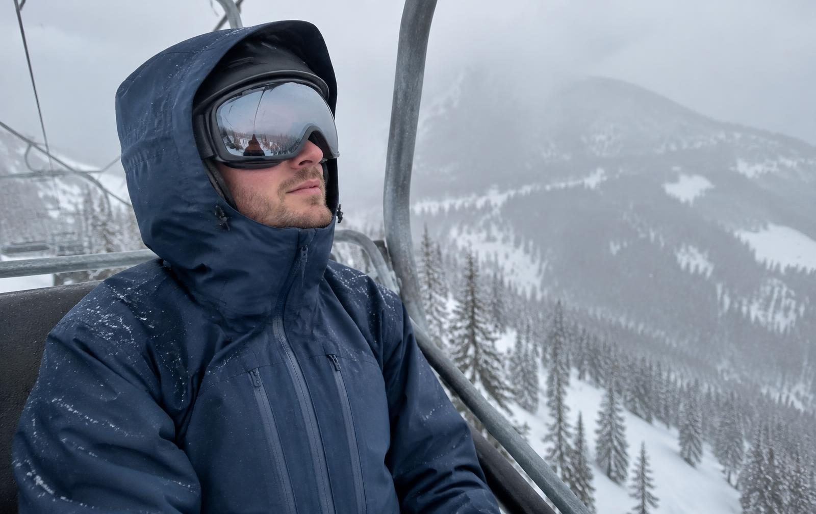 A man in a navy insulated ski jacket with the hood up riding a chairlift in falling snow