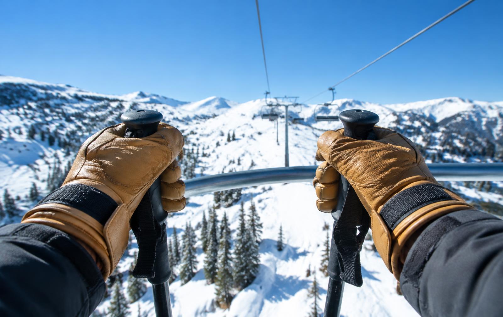 A skier's hands in leather ski gloves holding poles on a chairlift above snowy trees