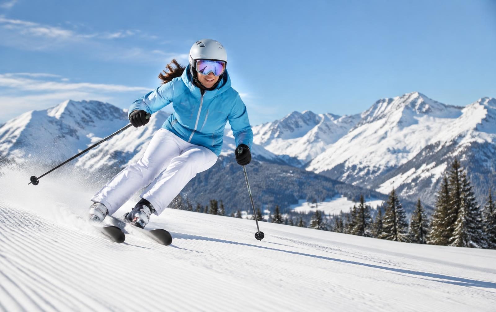 A woman in a light blue ski jacket carving a turn on a freshly groomed run below snowy peaks